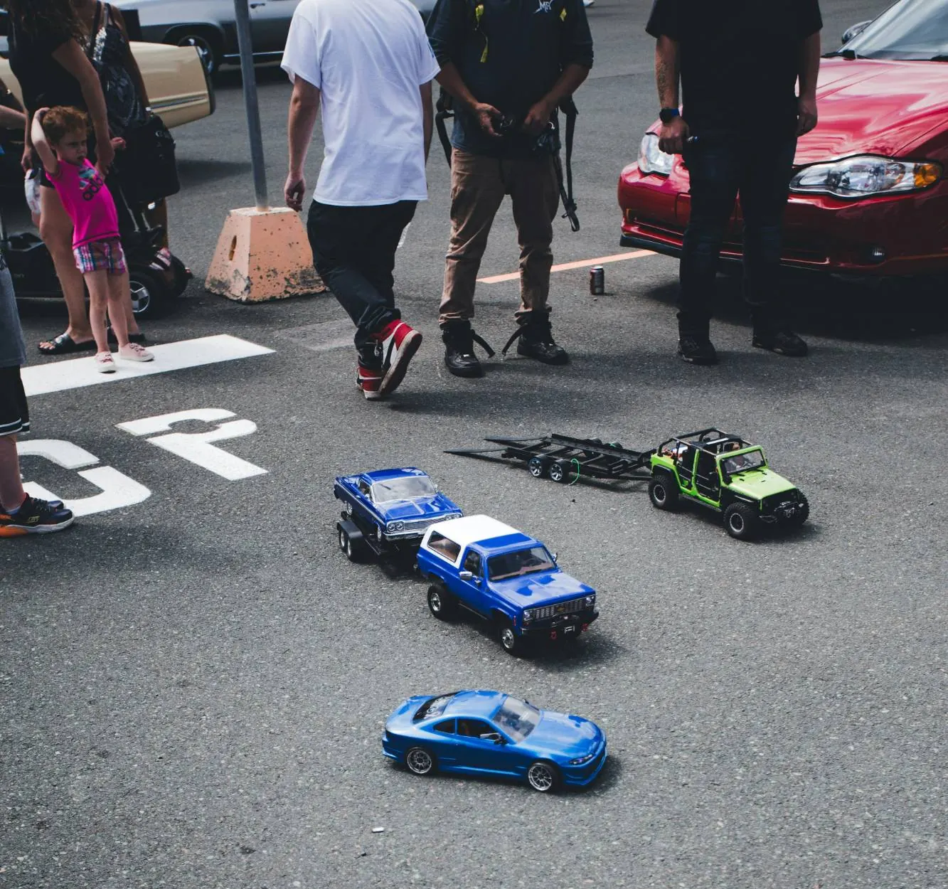 RC cars lined up on the tarmac at a meetup: a blue drift car, a blue truck towing a trailer and a green crawler